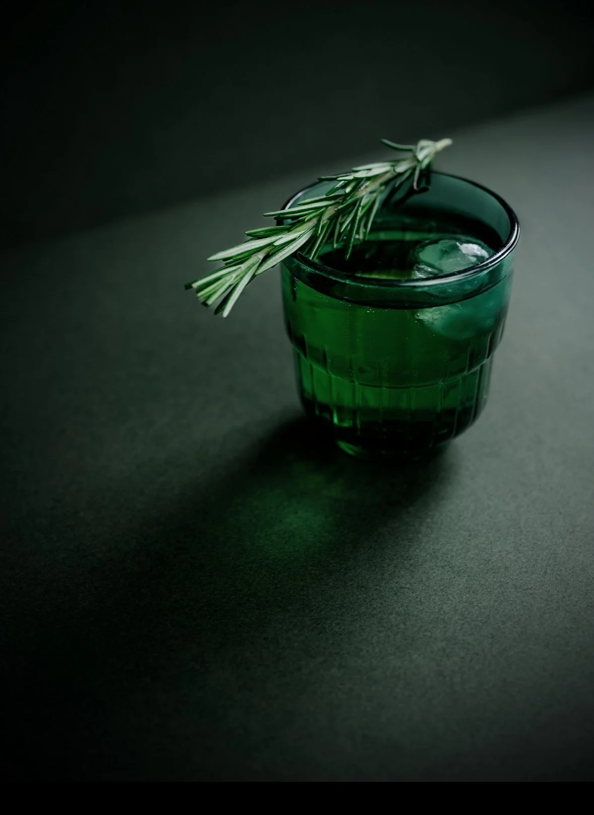Verre vert avec une branche de romarin dessus, placé sur une surface sombre.