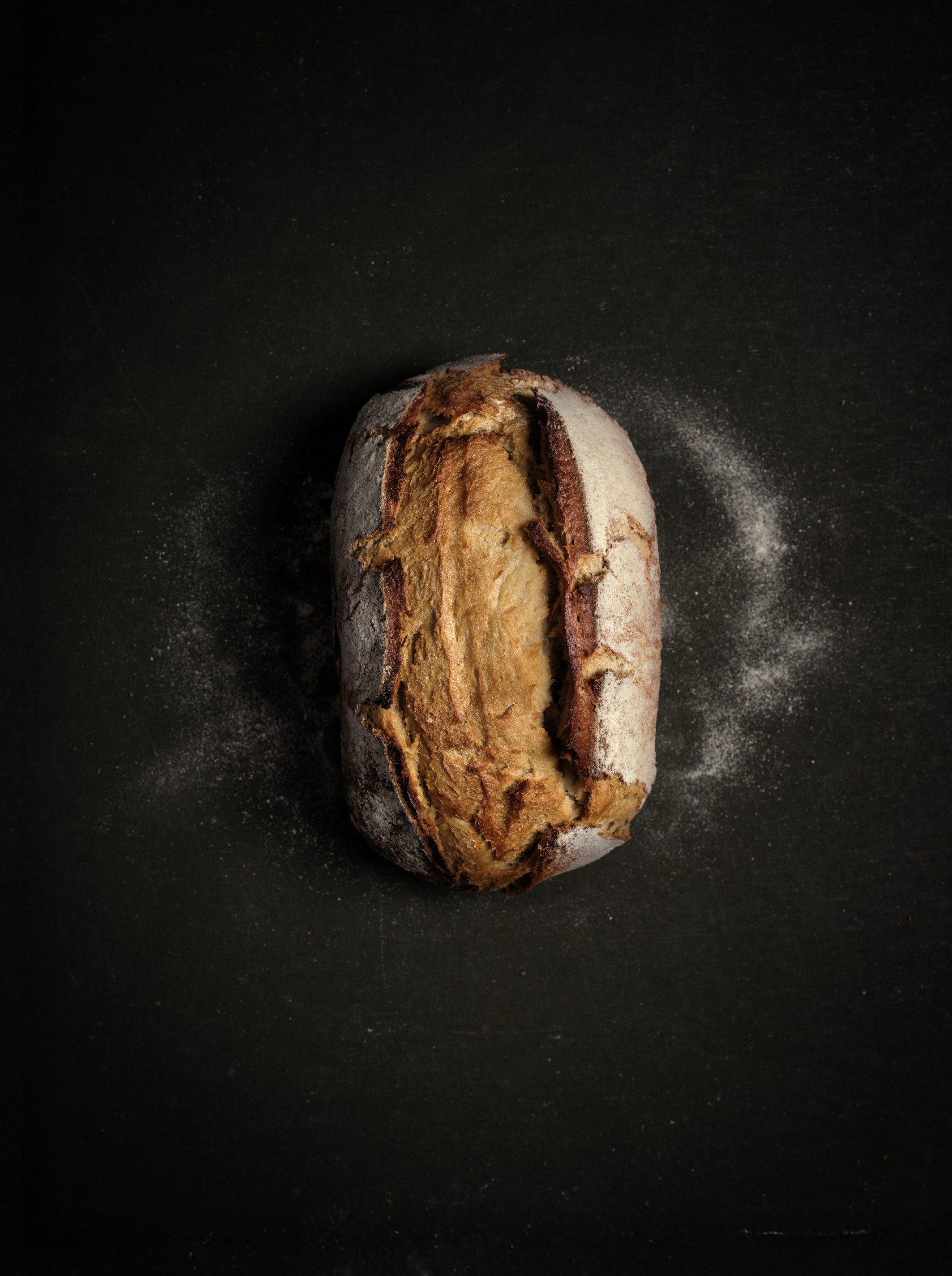 Pain artisanal posé sur un fond sombre, éclairage doux mettant en valeur la croûte et la farine dispersée autour.