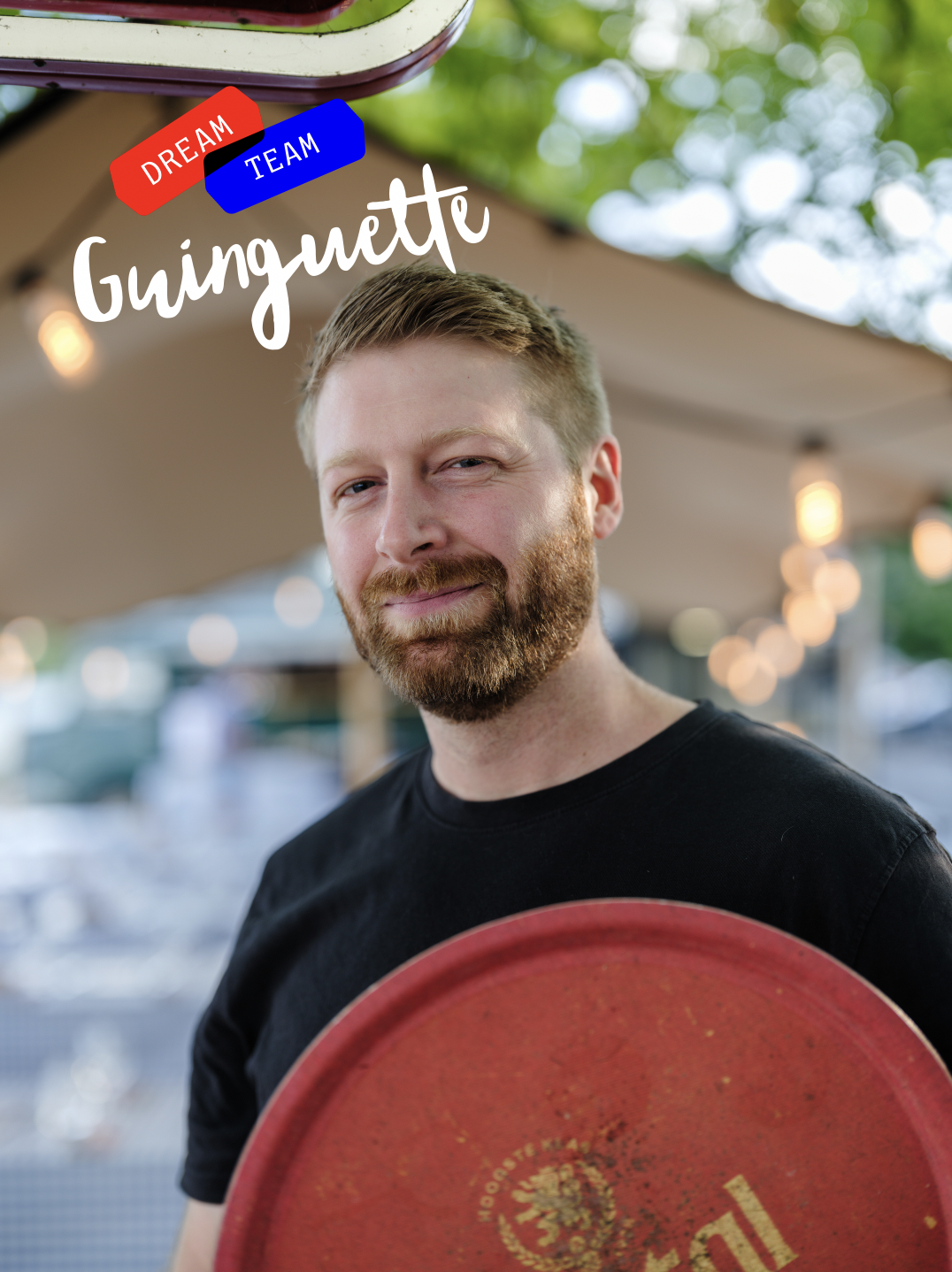 Un homme à barbe souriant, tenant une grande assiette rouge, en plein air avec des décorations lumineuses et des arbres en arrière-plan.