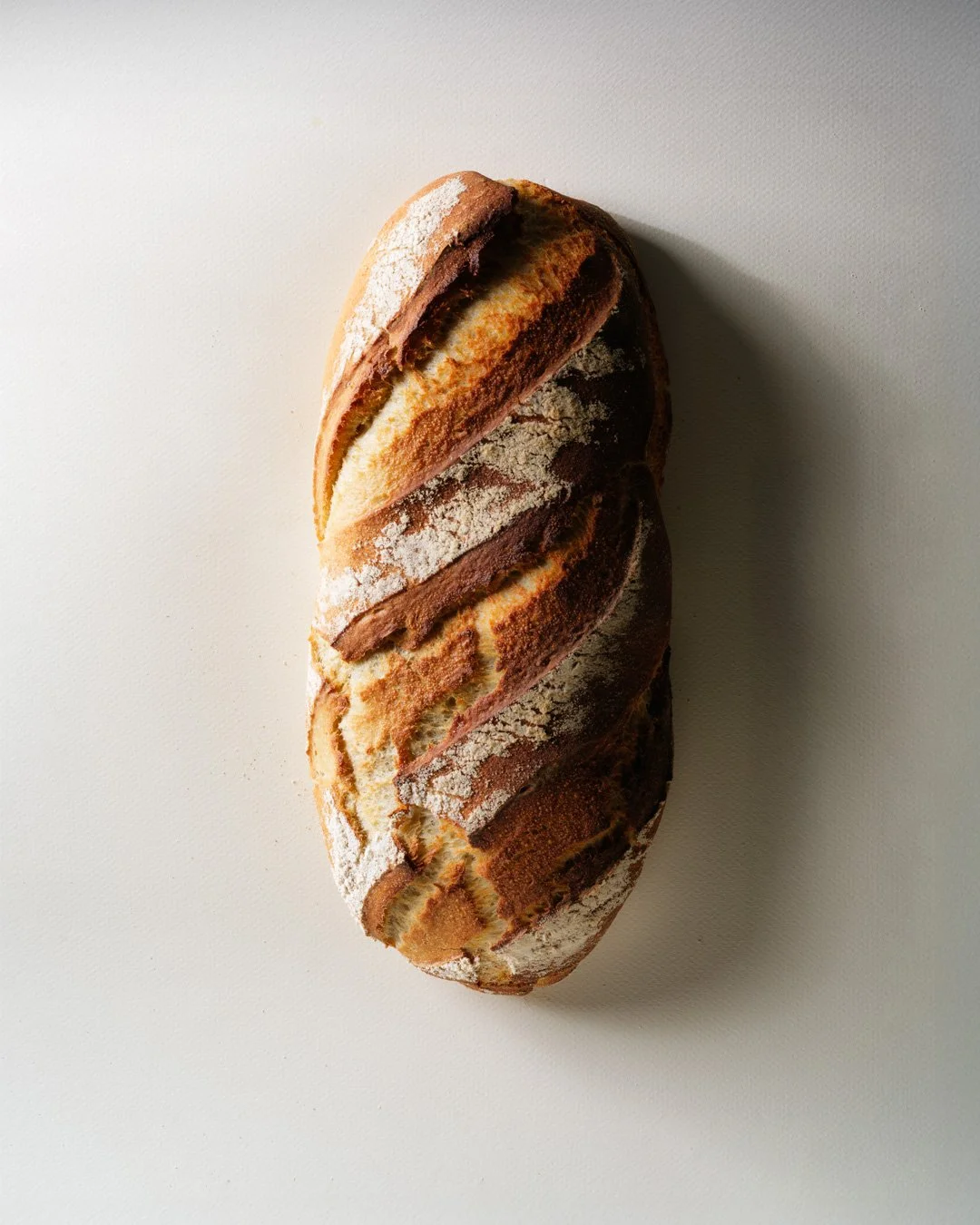 Pain artisanal rectangulaire avec une croûte dorée et légèrement brûlée, saupoudré de farine, posé sur une surface blanche.
