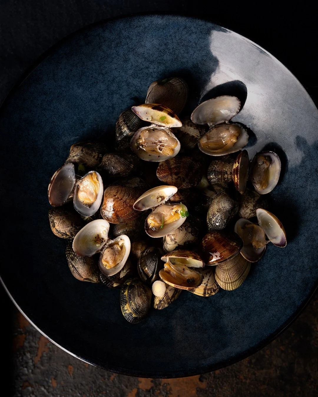 Assiette bleu pétrole remplie de palourdes ouvertes, éclairée en lumière sombre et directionnelle. Textures brillantes, plateau métallique rouillé en dessous, ambiance dramatique et éditoriale.