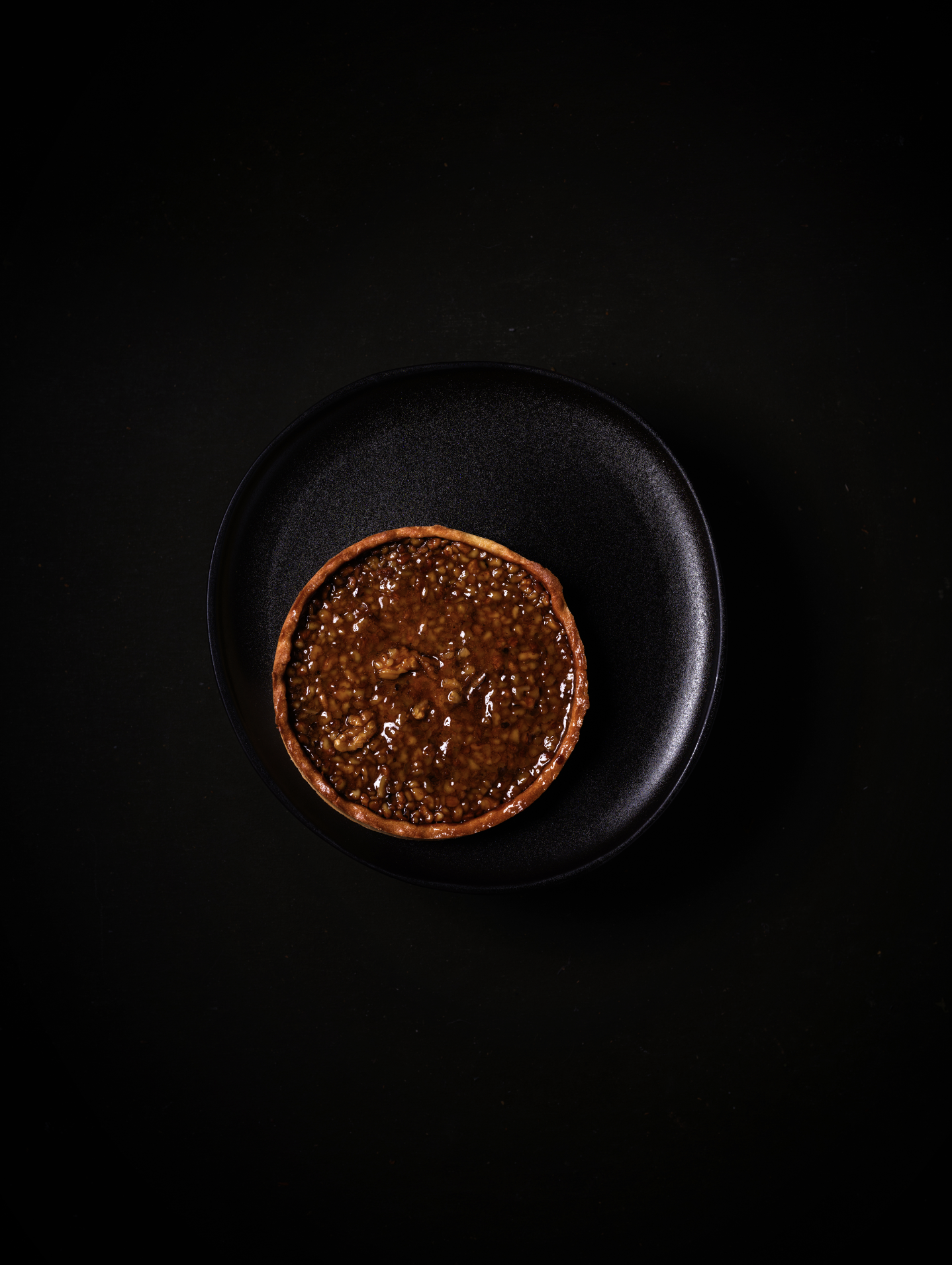 Tarte aux noix photographiée en vue du dessus sur une assiette noire, éclairage sombre et minimaliste.