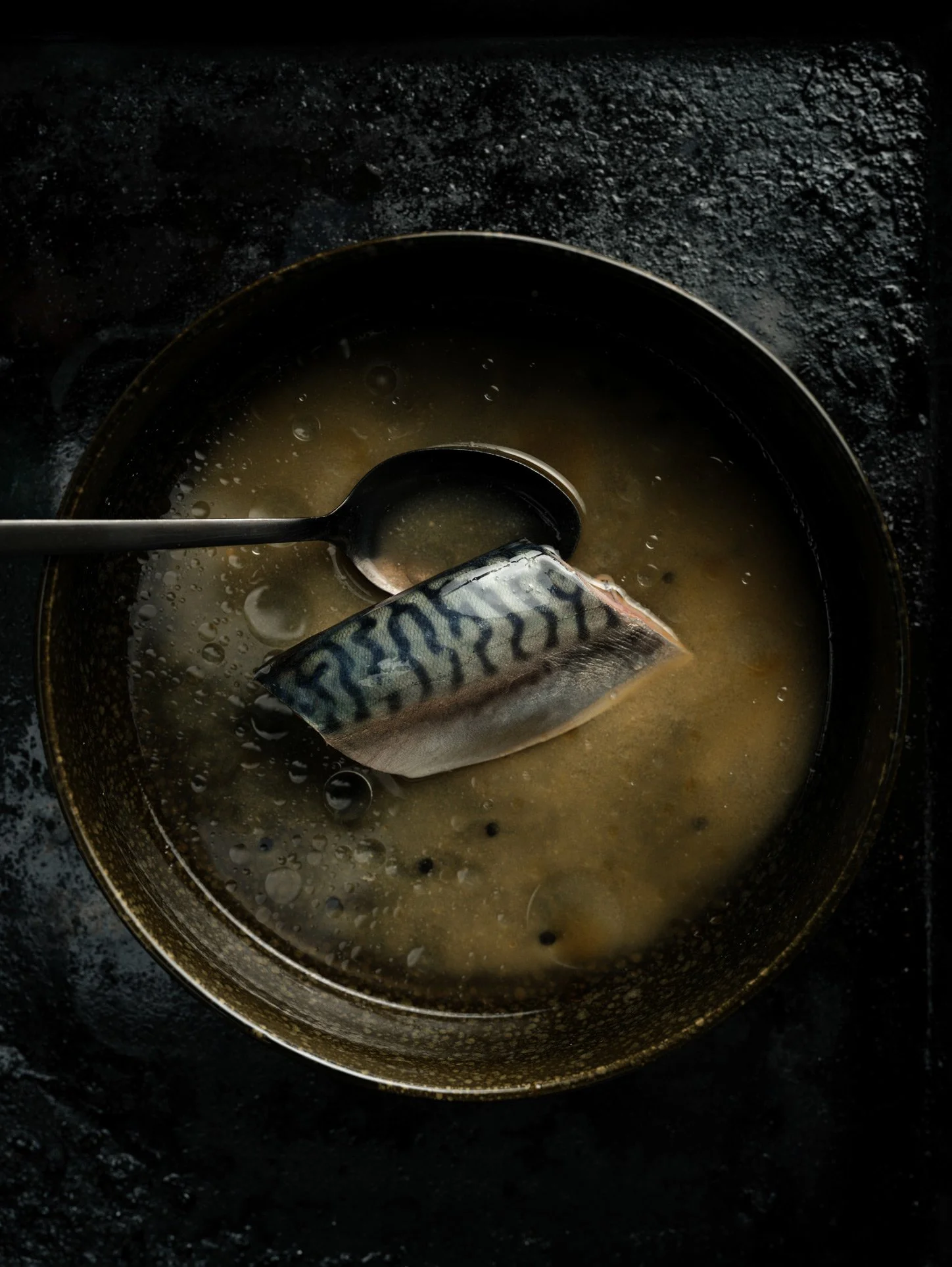 Filet de maquereau cru dans un bouillon ambré, photographié en vue du dessus dans un bol sombre sur fond noir texturé, avec une cuillère en métal.