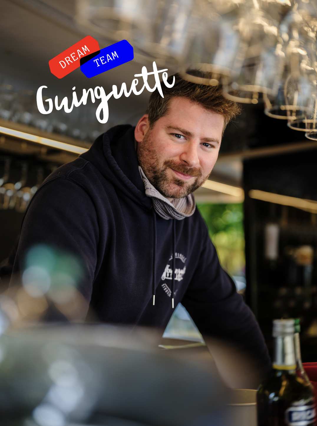Un homme souriant dans un café ou restaurant, portant une veste à capuche noire, avec des verres suspendus au plafond et des bouteilles sur le comptoir. Texte en français : "Ginguette, Dream Team".