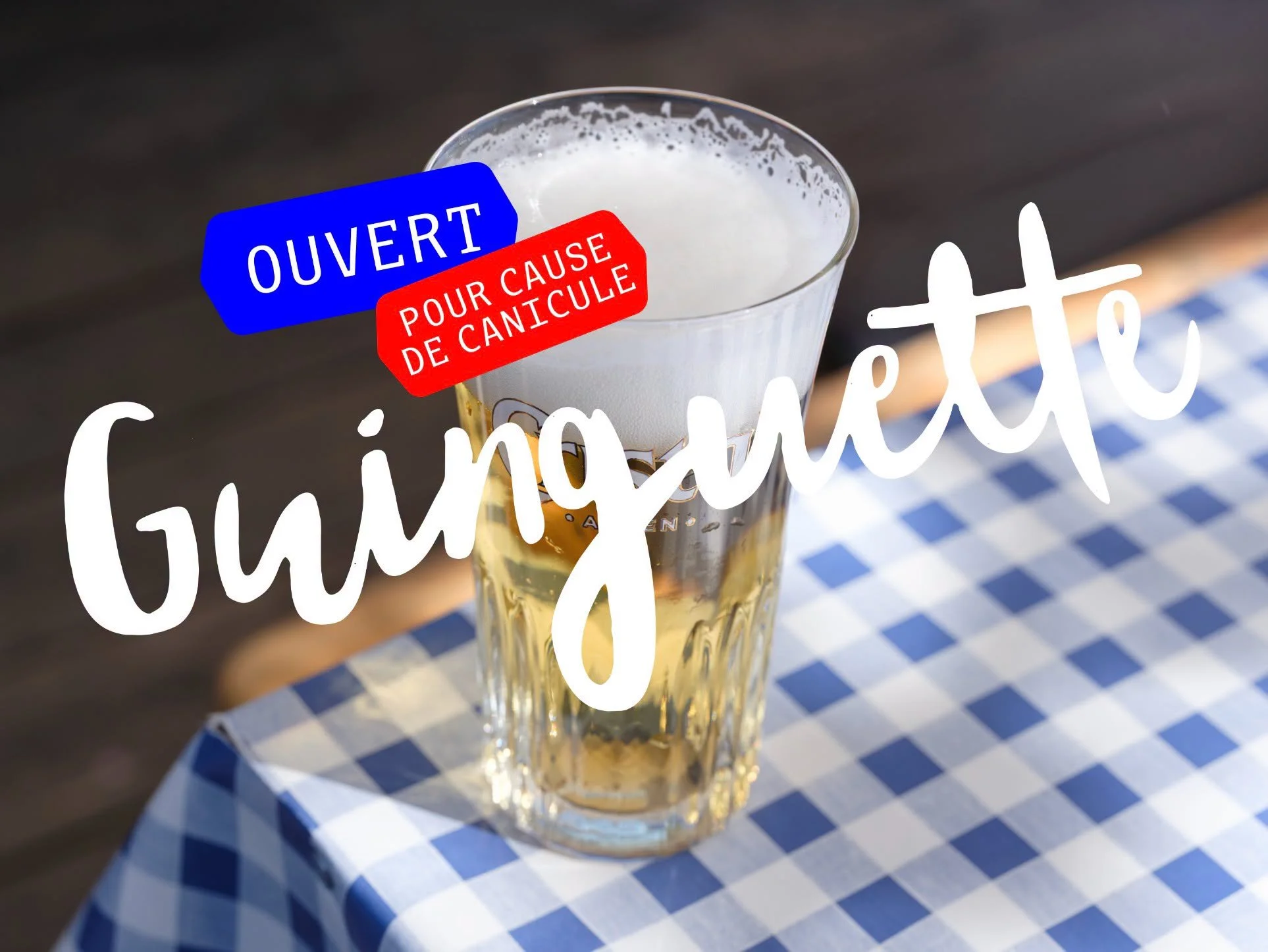 Verre de bière avec un label bleu et rouge indiquant 'OUVERT POUR CAUSE DE CANICULE' sur une table à carreaux bleus et blancs.