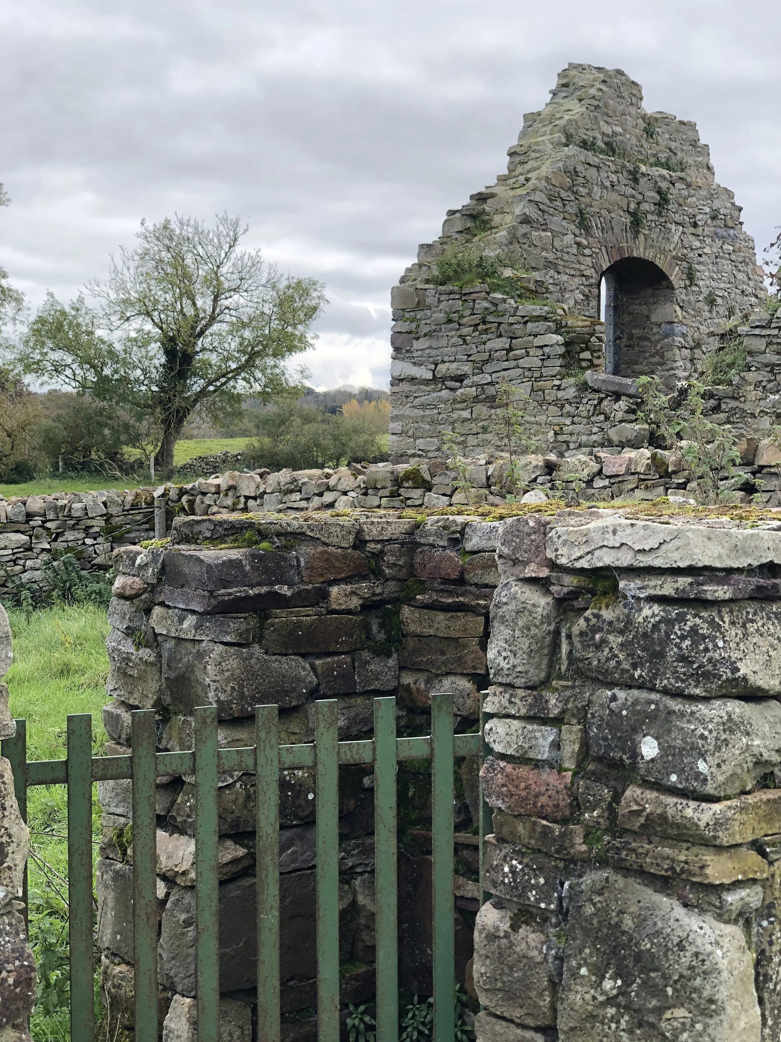 Ruins of an old stone building with arched window, surrounded by stone walls and metal fence, with a tree and cloudy sky in the background.
