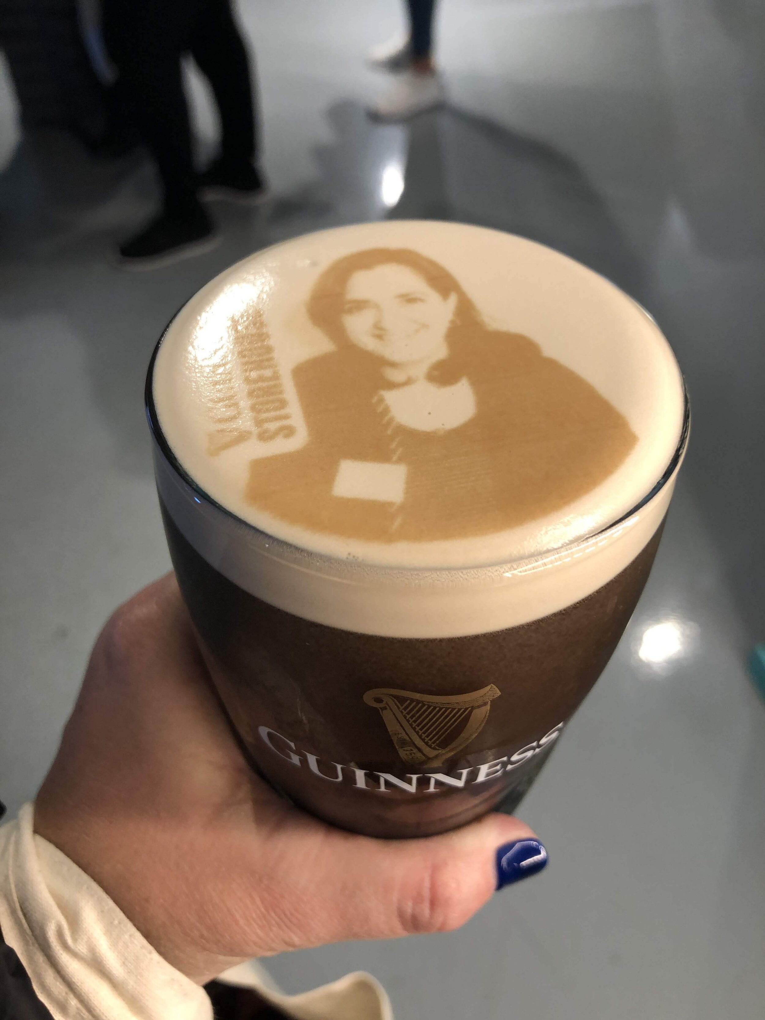 A pint of Guinness beer with latte art featuring a portrait of a woman and the word 'SUE' on the foam.