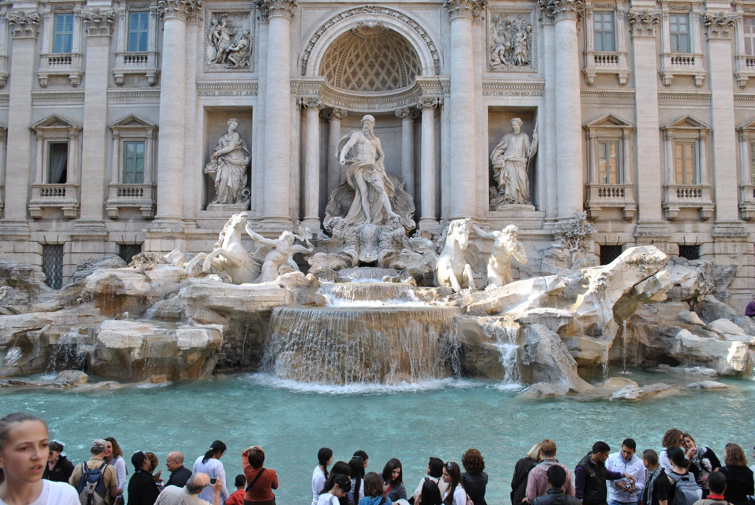 The Trevi Fountain in Rome, Italy, featuring intricate baroque sculptures of gods and mythological figures, with water flowing into a large basin surrounded by tourists.