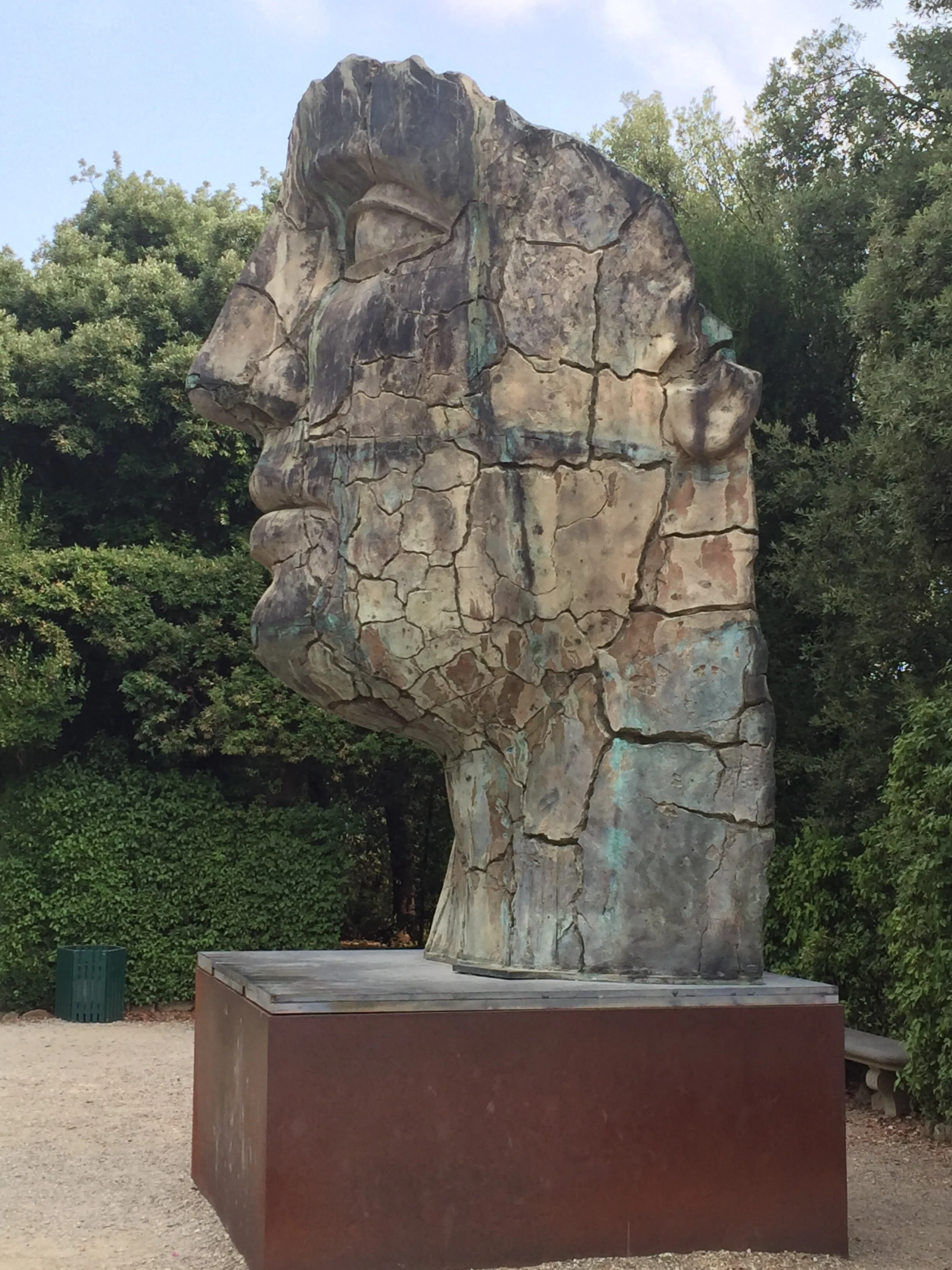 A large sculpture of a human face in profile, made of cracked stone or concrete, placed on a rectangular base outdoors. Surrounding trees and bushes are visible in the background.