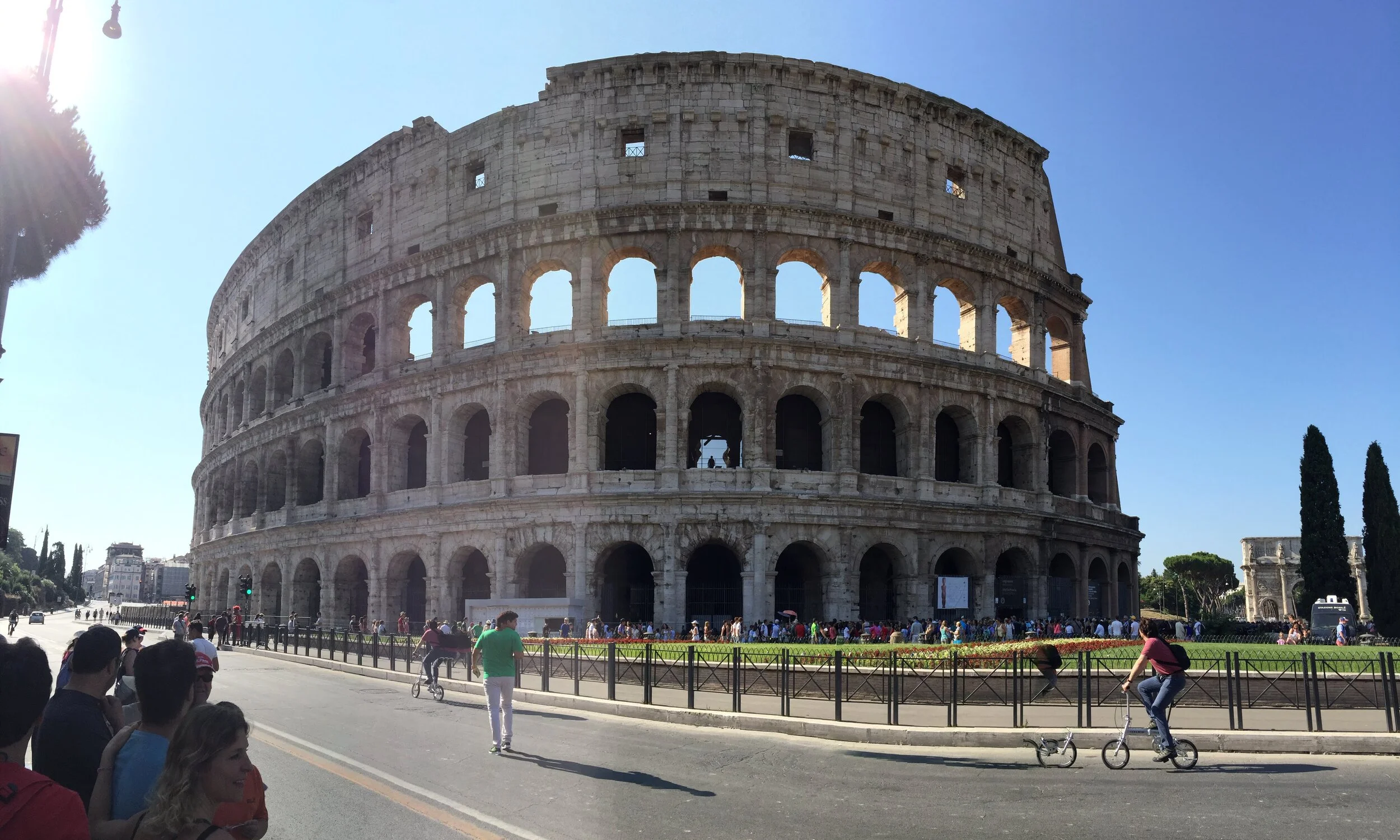 The Colosseum in Rome, Italy, with tourists and people riding bikes in front, under a clear blue sky.