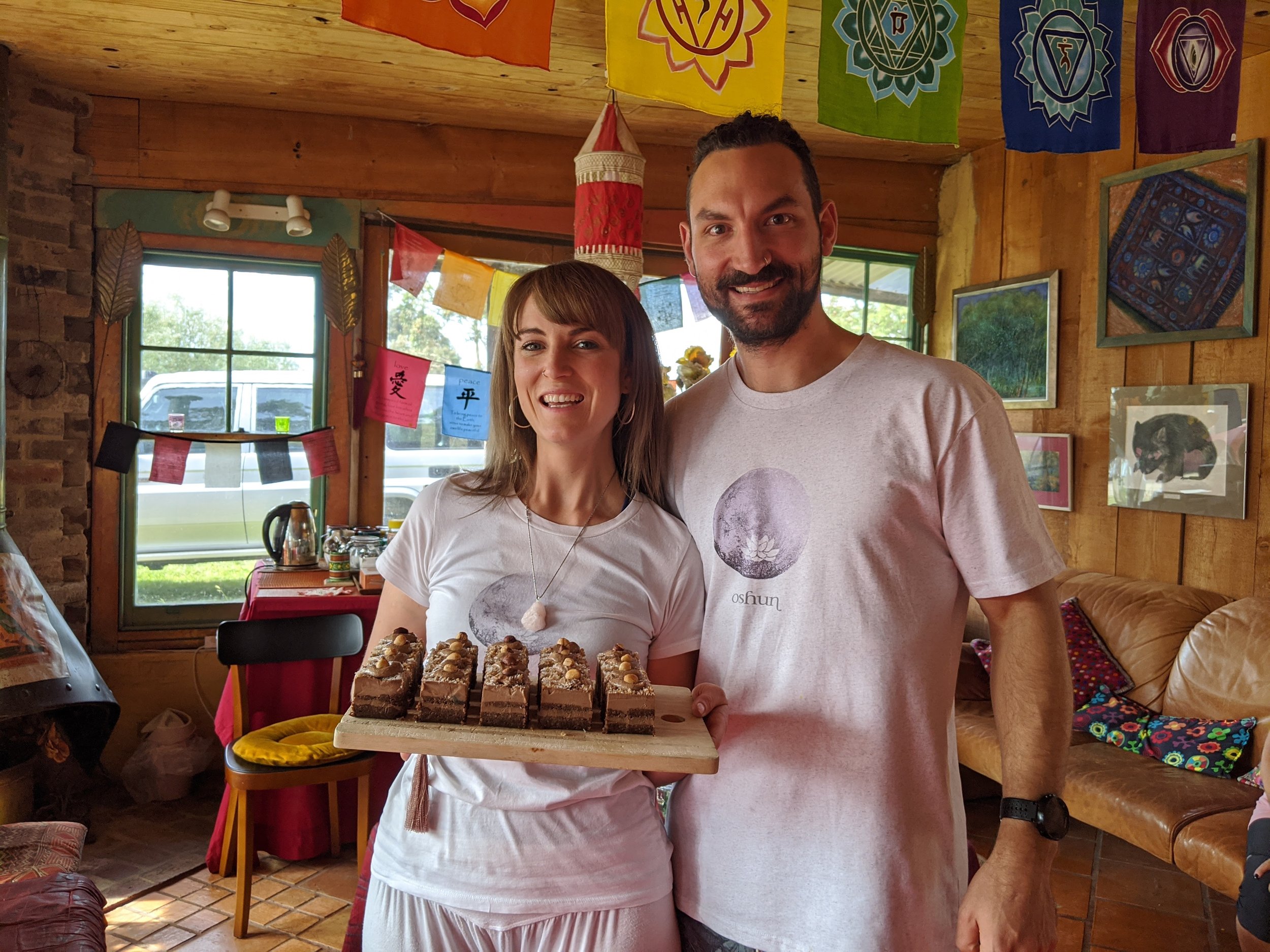 Two people smiling indoors, one holding a tray of desserts. The room has colorful decorations and banners with symbols on the walls.