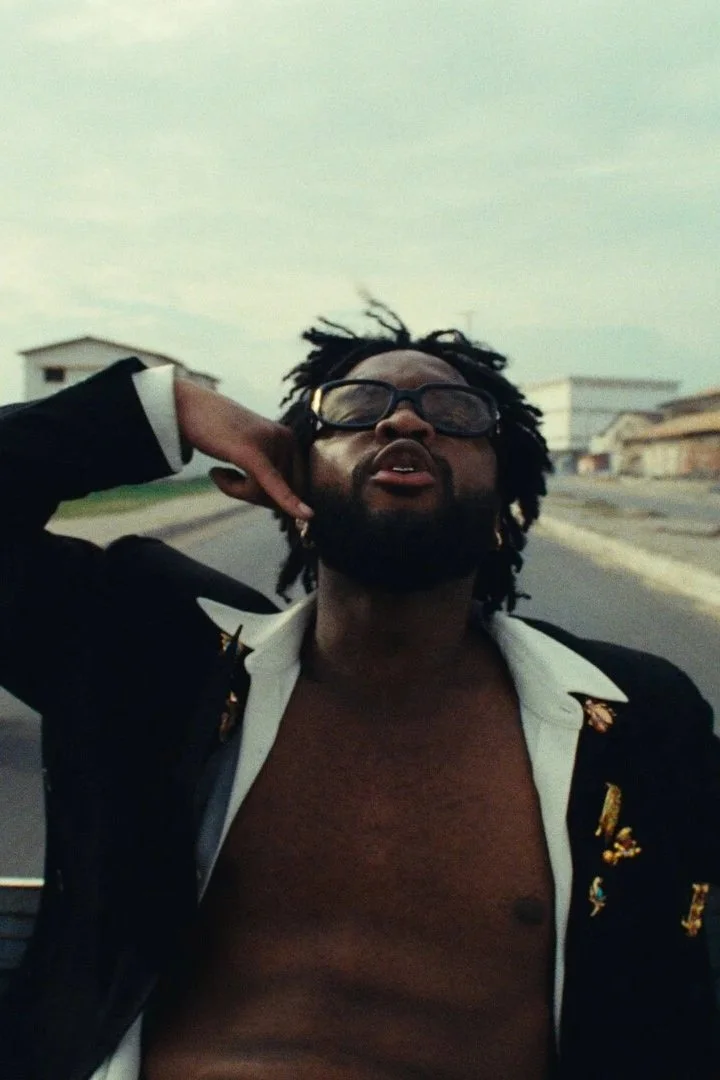 A man in a suit jacket and sunglasses sitting on the back of a truck, mid-song.