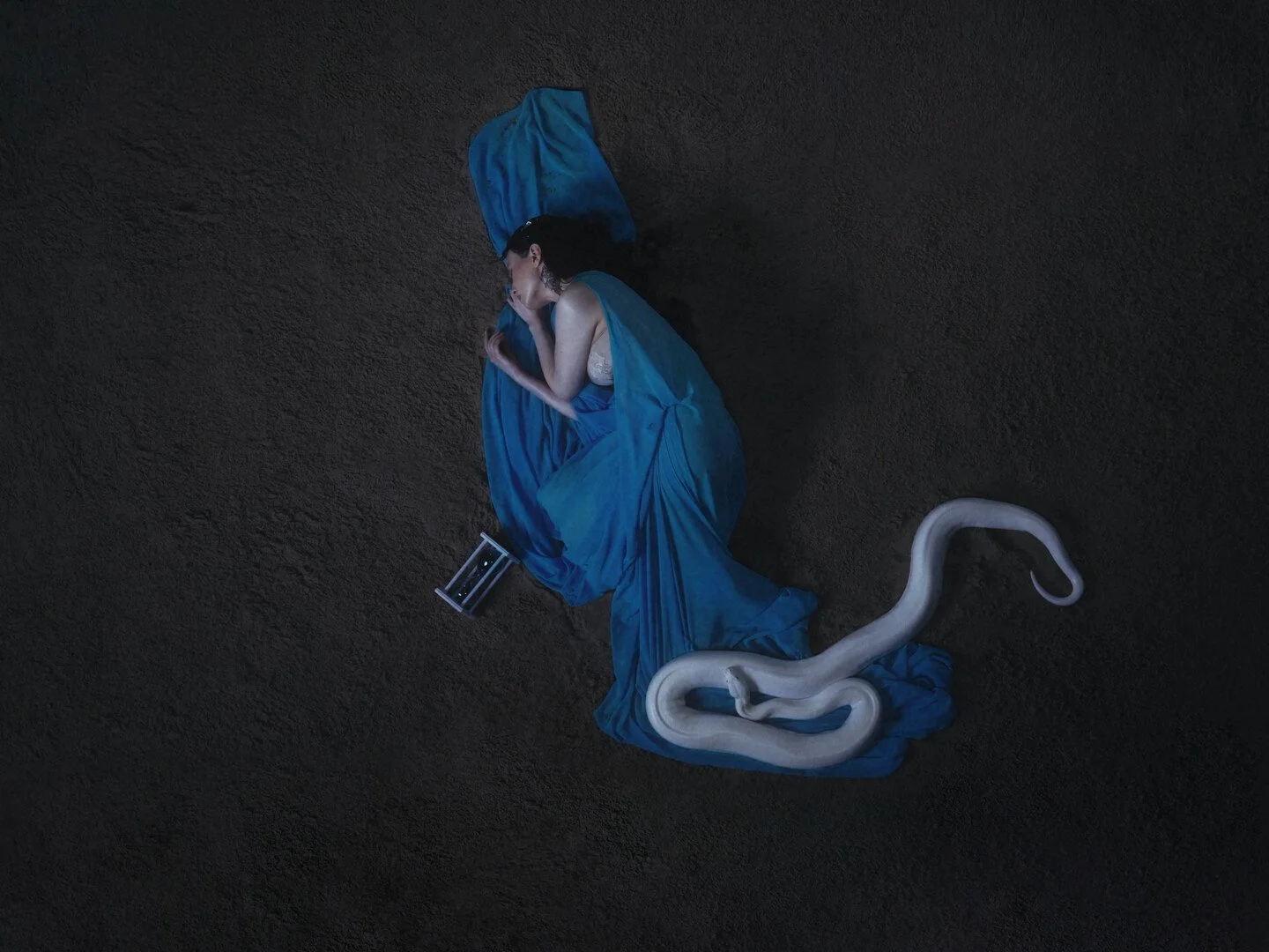 A woman in a flowing, blue dress, laying on the desert floor with a snake at her feet.