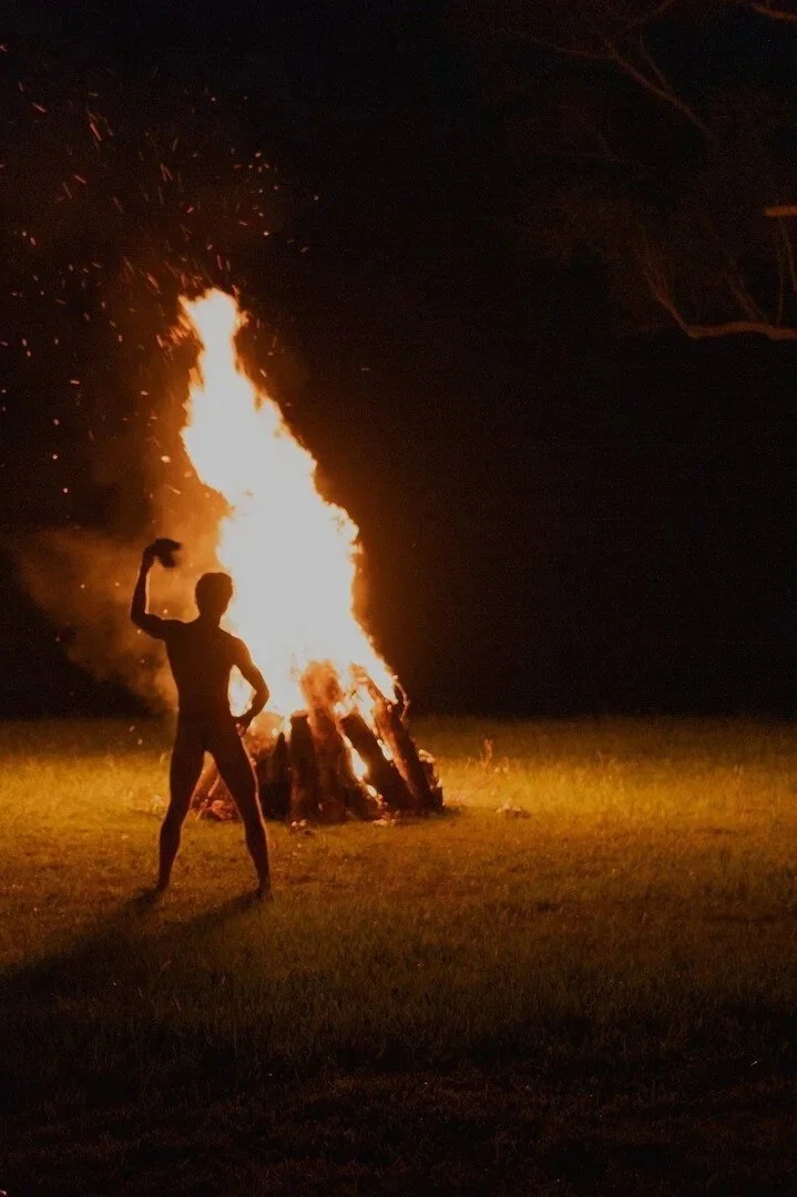 A man standing by a large, blazing fire pit waving around a piece of clothing.