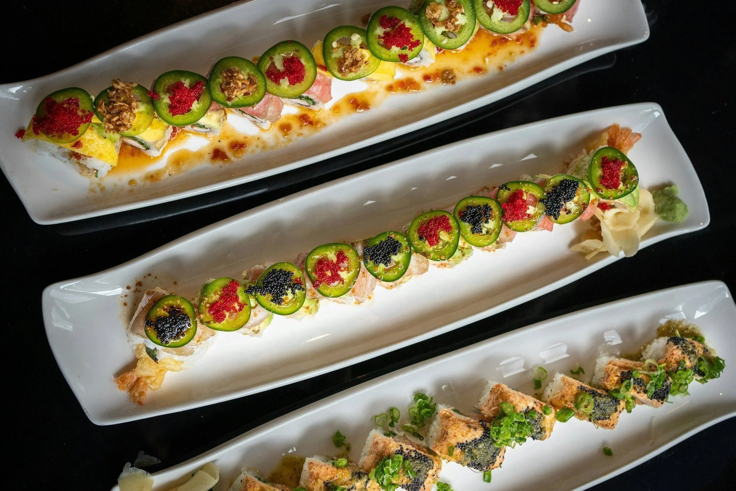 Three long white rectangular plates with assorted sushi rolls, garnished with sliced jalapeños, fish roe, and sauce, placed on a black surface.