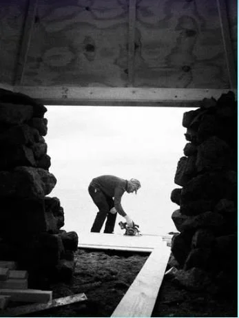 A person wearing a cap is bending over outdoors, working on a plank of wood with a tool. Framed by a dark stone doorway, the tone is industrious and focused.