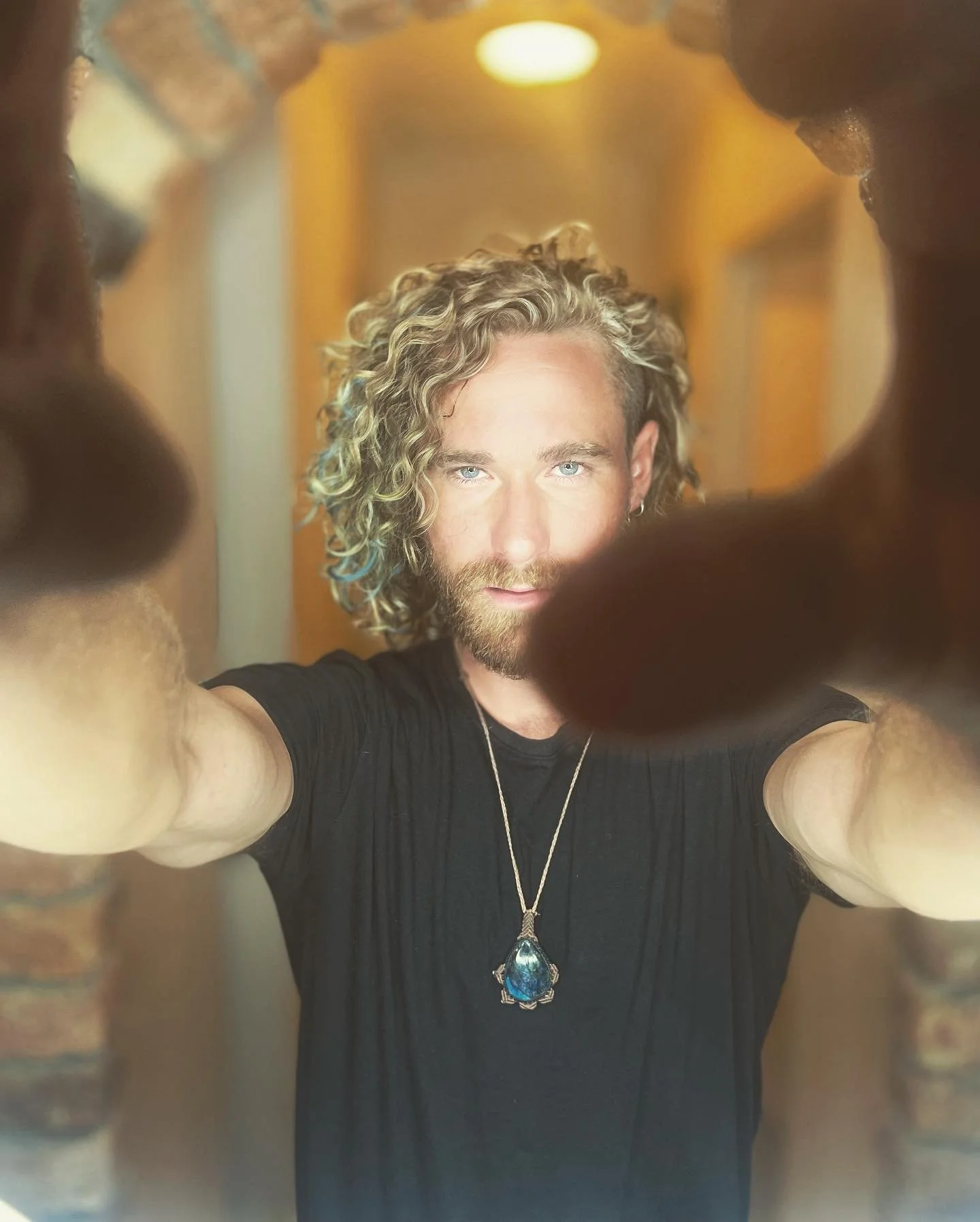 A man with curly blonde hair and a beard taking a selfie through a small opening, wearing a black shirt and a large blue pendant necklace.