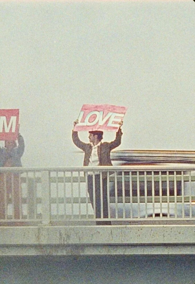 A man standing on a highway bridge with a painted sign that reads ' LOVE'.