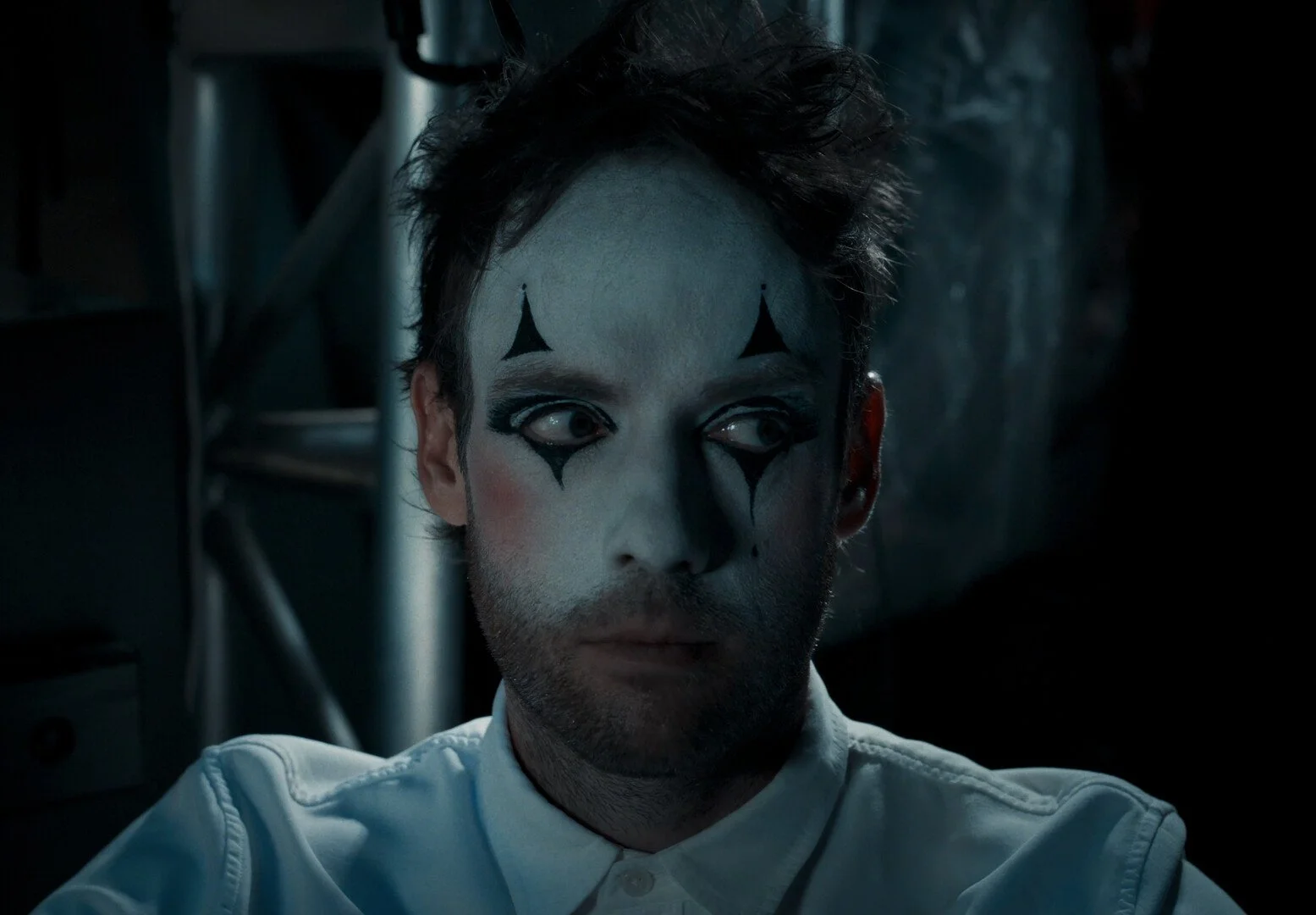 A man in marionette makeup, with a short beard, his hair askew and looking to his left.