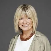 A professional headshot of a smiling woman with short blonde hair, wearing a light jacket and a red necklace against a neutral background.