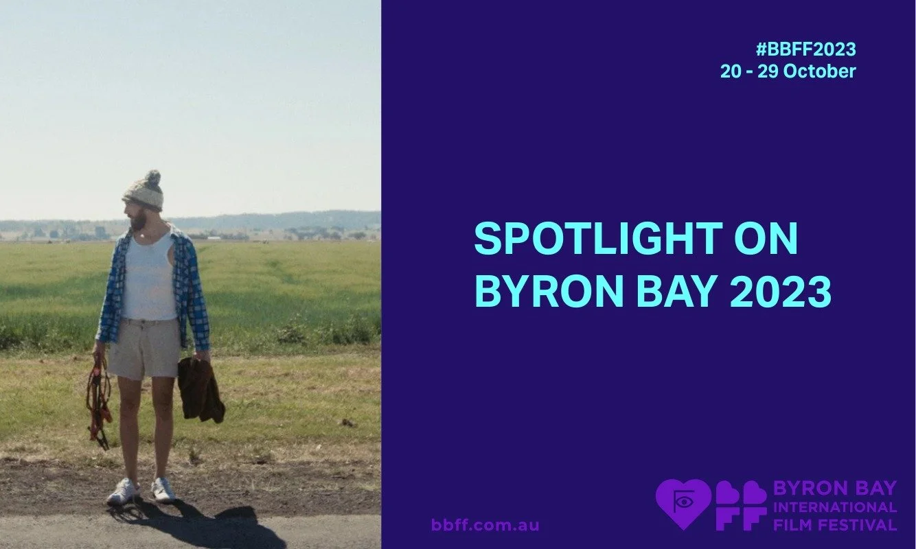 Person in casual attire stands on a rural road with fields, wearing a beanie and shorts, holding a bag. Blue poster reads "Spotlight on Byron Bay 2023."