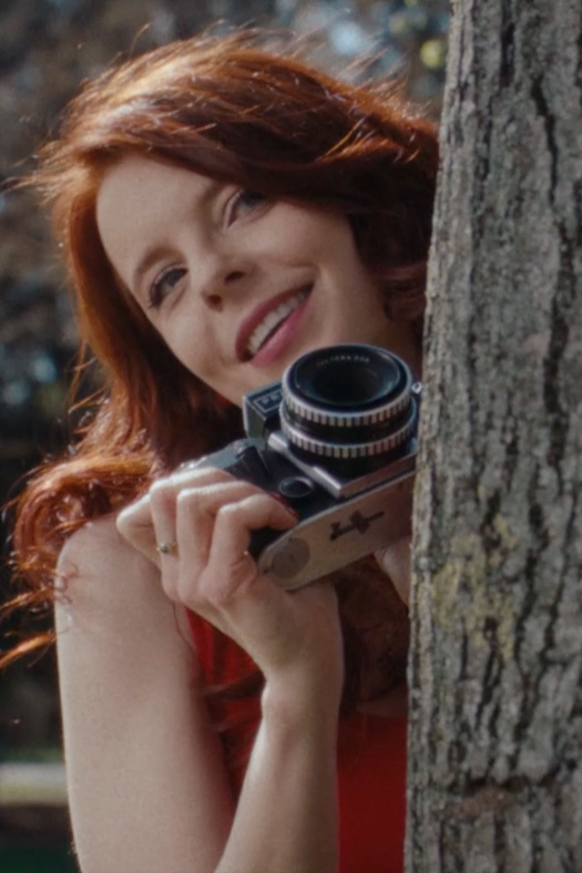 A woman with red hair hiding behind a park tree with a camera.