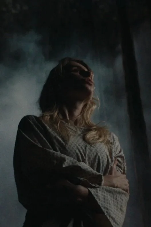 A woman with long, messy blonde hair hugging her hospital gown close to her chest.