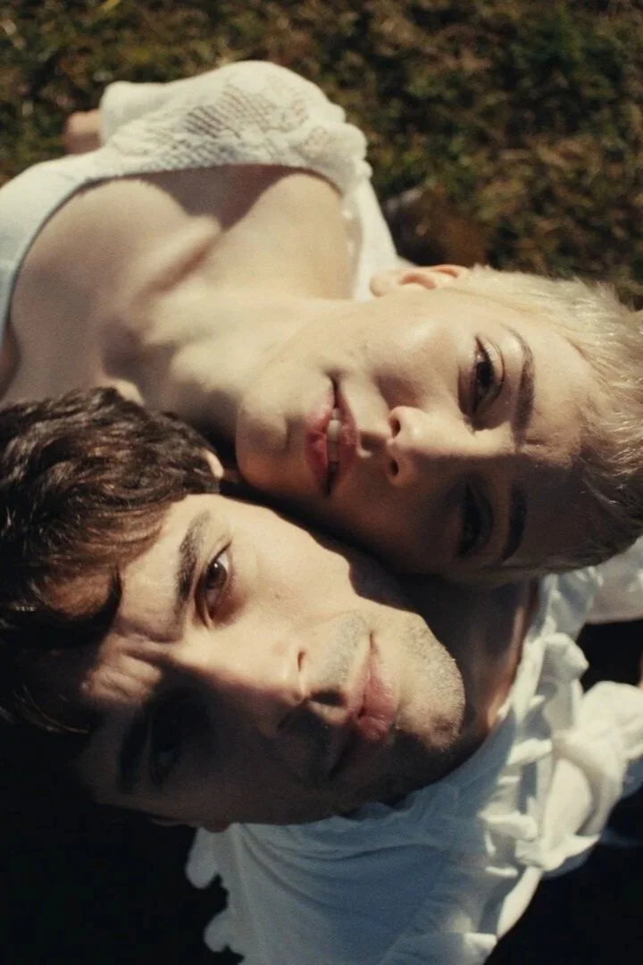 A woman with short bleach blonde hair and a man with short brown hair standing in a field with the side of their heads pressed against on another.