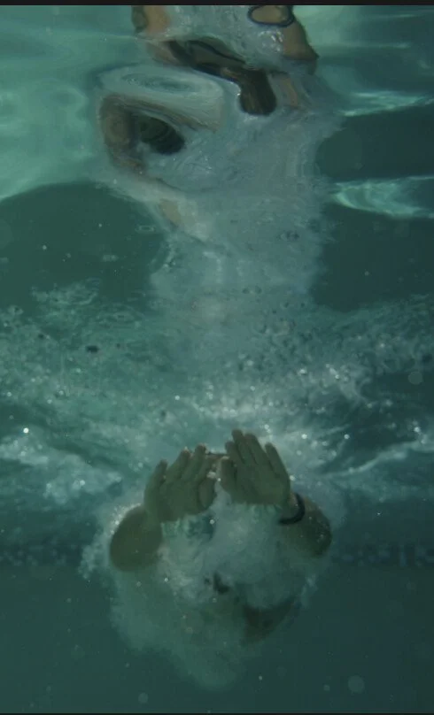 Underwater shot showing a person submerged face-down, hands raised near their head amid a swirl of bubbles, with a blurred reflection above the water's surface.