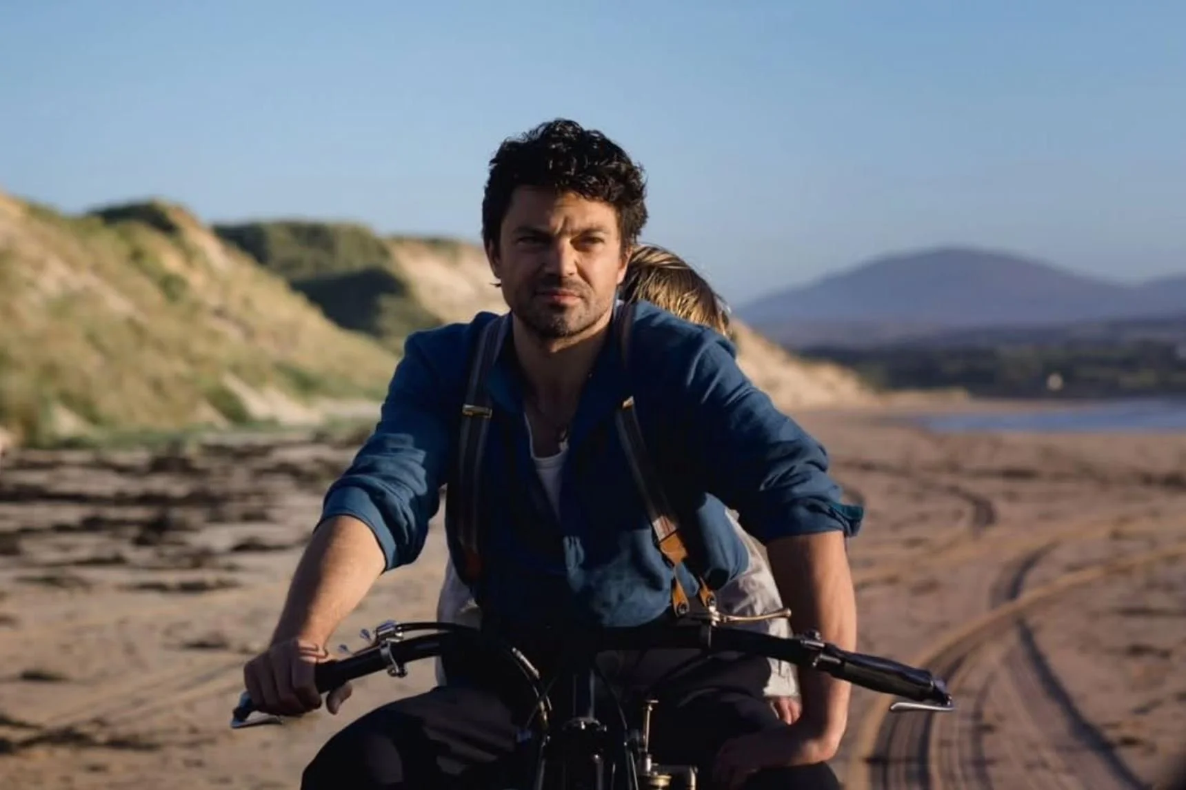 Film still from The Lightkeeper. A man rides a bicycle along a sandy beach at sunset, a woman seated behind him with her head resting on his back, coastal hills in the background.