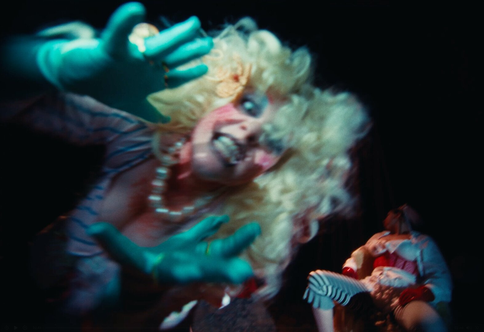 A woman with curly blonde hair, dressed like a doll with doll makeup grabbing at the camera. with a crazed look on her face