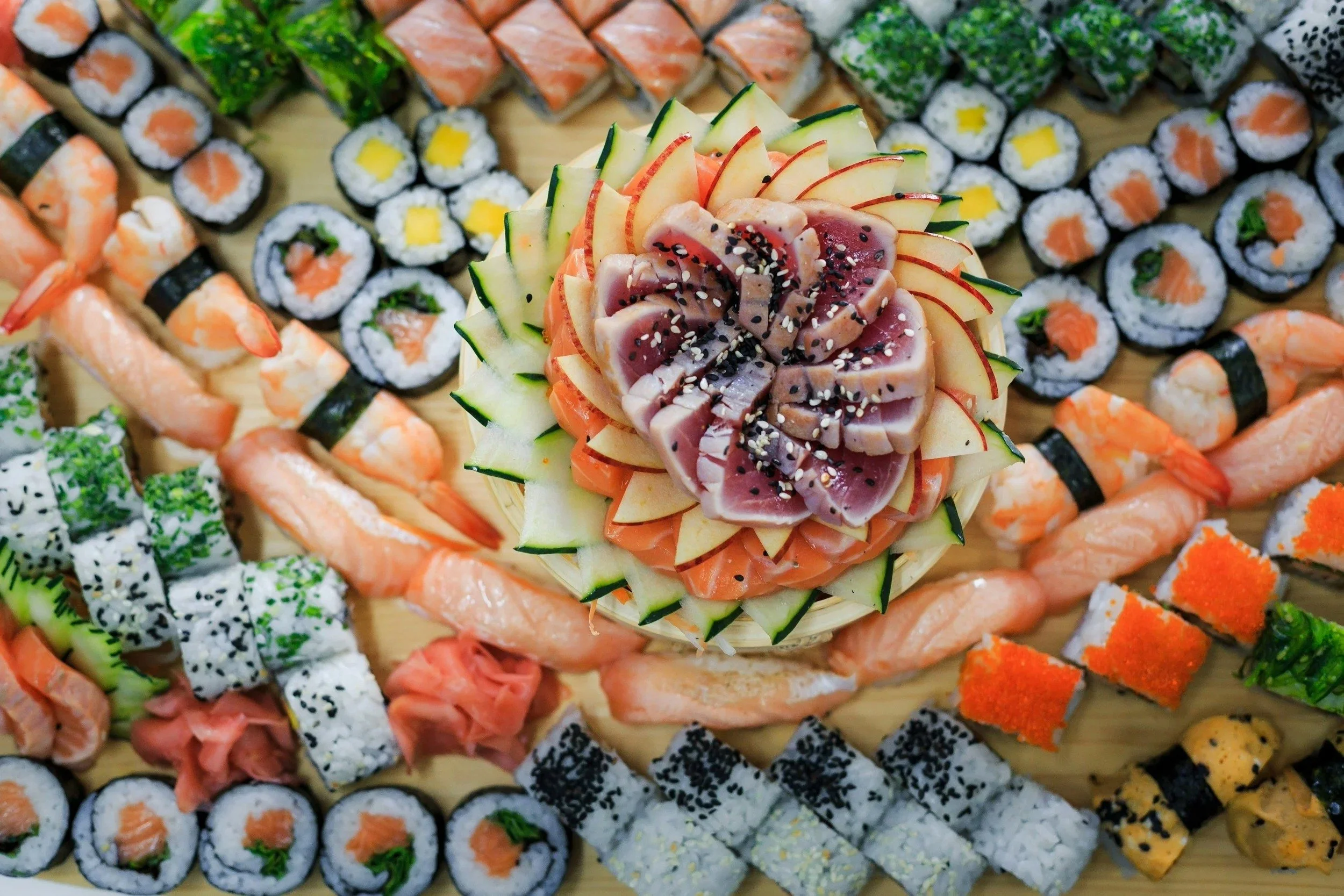 Assorted sushi rolls surrounding a decorative platter with sliced raw tuna, salmon, and cucumber garnishes.