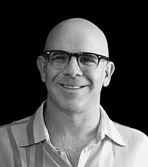 A black-and-white headshot of a smiling bald man wearing glasses and a striped shirt against a dark background.