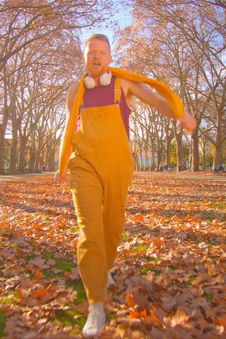 A man in a bright yellow overalls, yellow scarf and a magenta singlet walking through a park in autumn, mid-dance.