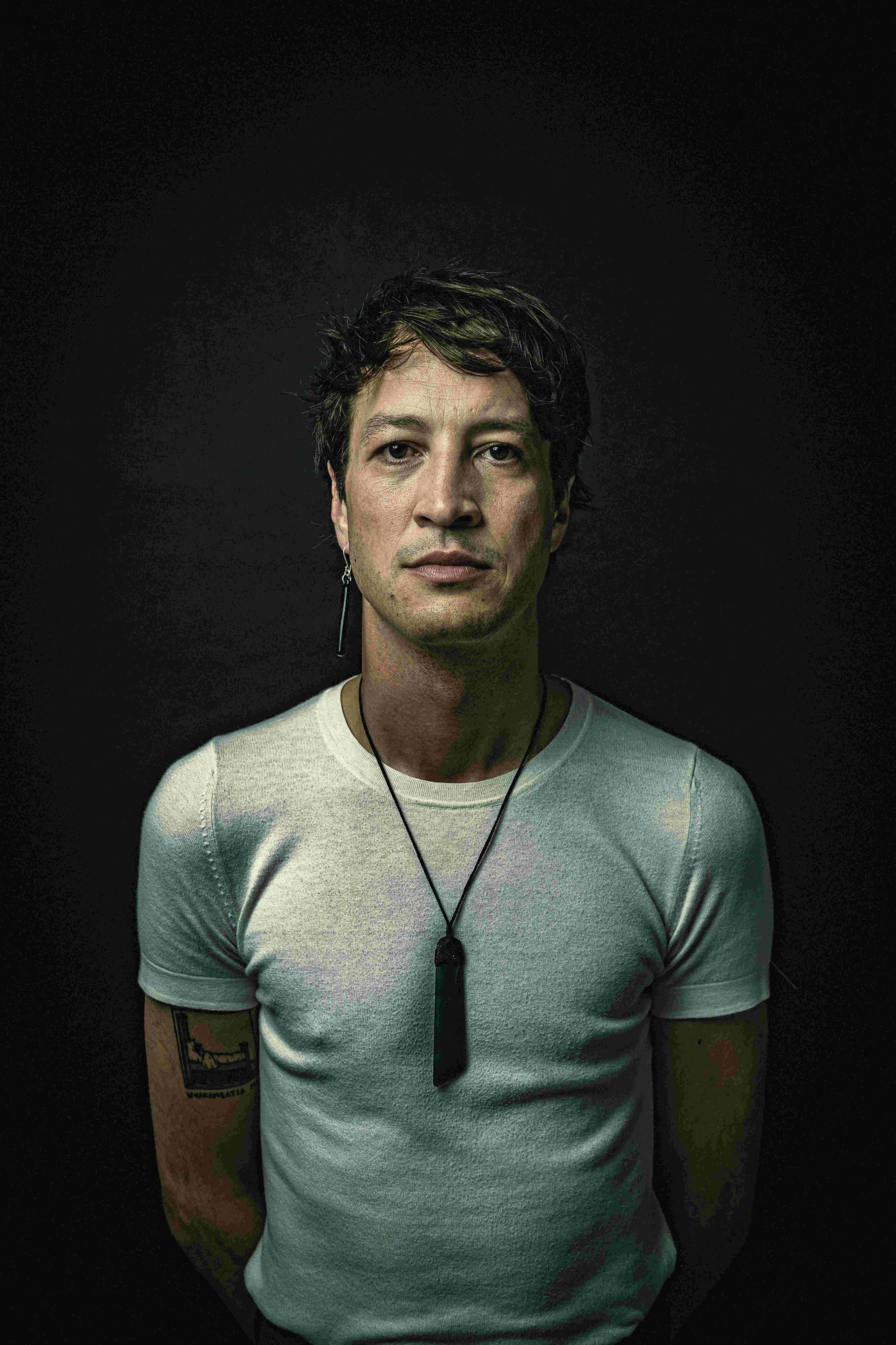 A man with short, dark, messy hair and an earring, wearing a white t-shirt and a black pendant necklace, standing against a dark background.