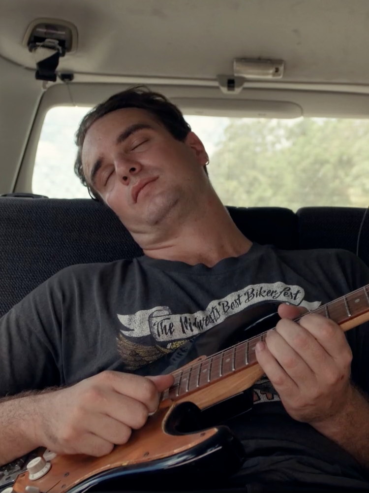 A man asleep in the backseat of a car with a guitar in his hand.