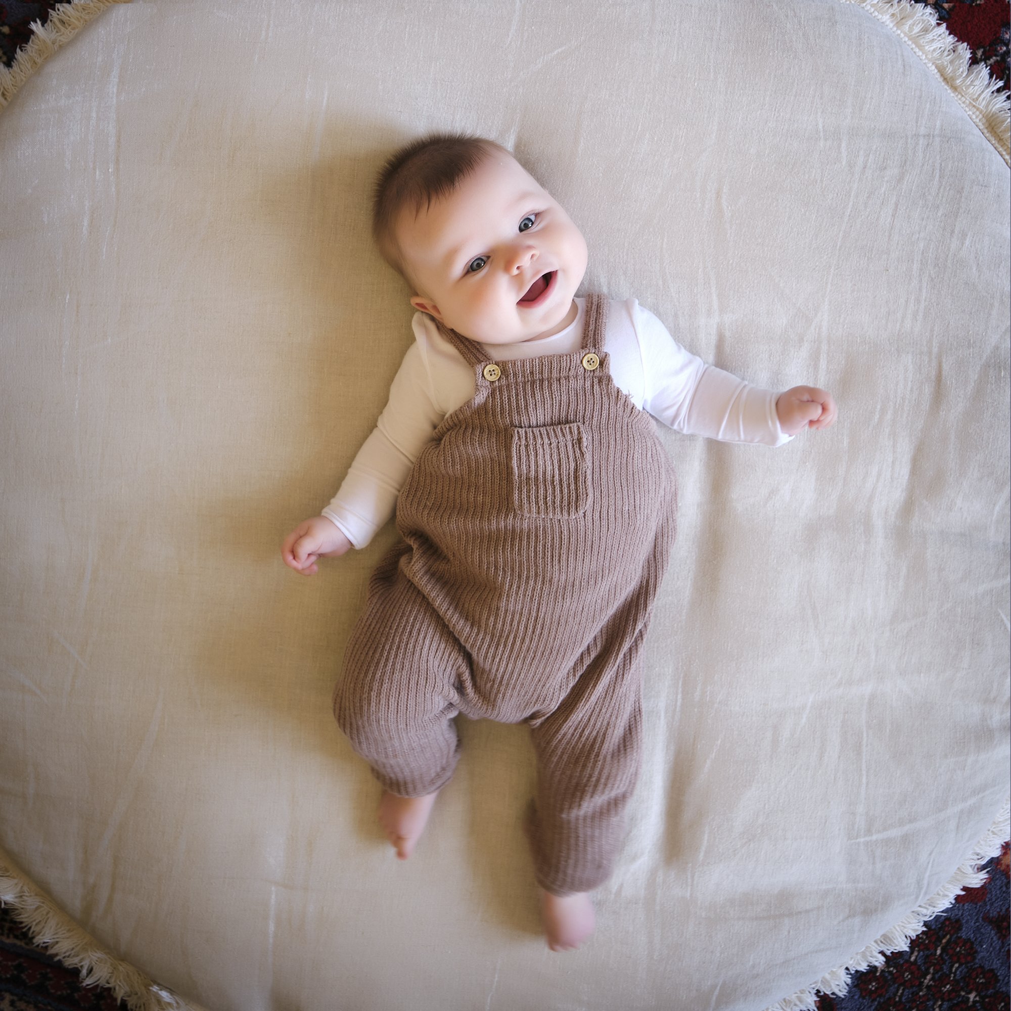 Smiling baby lying on a beige circular rug, wearing brown overalls and a white long-sleeve shirt.