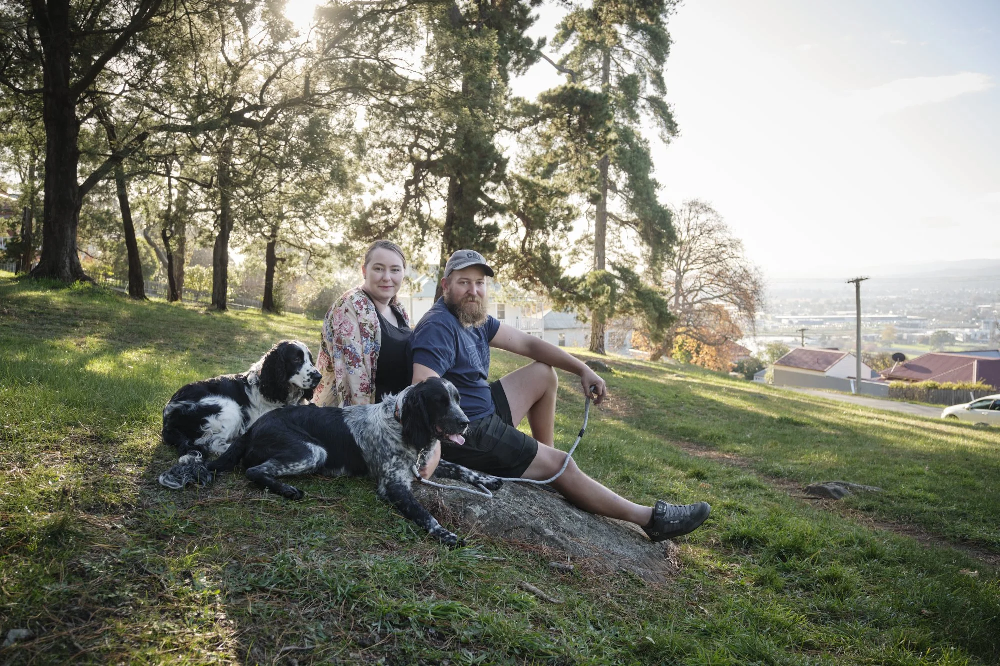 A couple sitting on a grassy hill with three black and white dogs, under large trees during late afternoon, overlooking a town with houses and power lines.