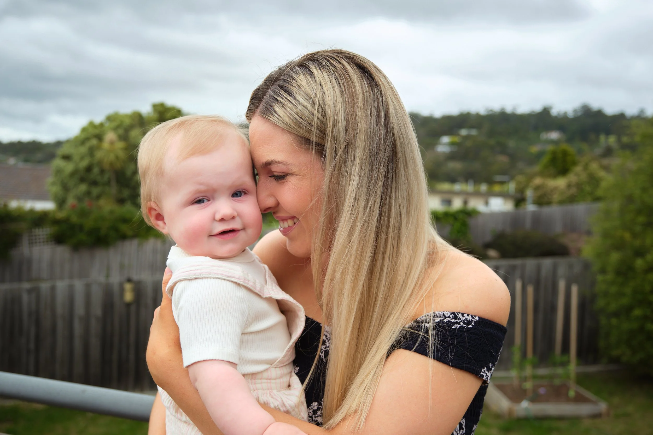A woman holding a young child outdoors on a cloudy day, with trees and a fence in the background. They are sharing a close, happy moment.