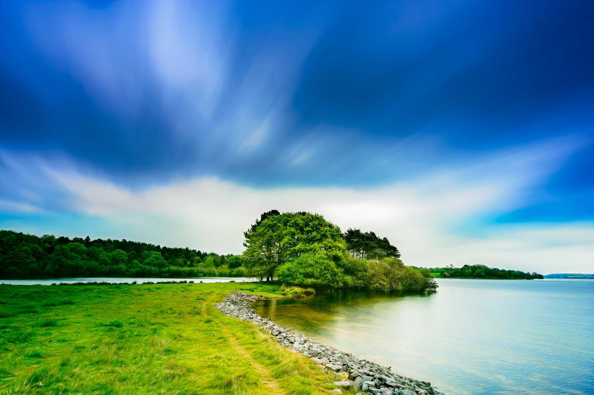 Grassy shoreline path at Rutland Water with trees and blurred sky from long exposure