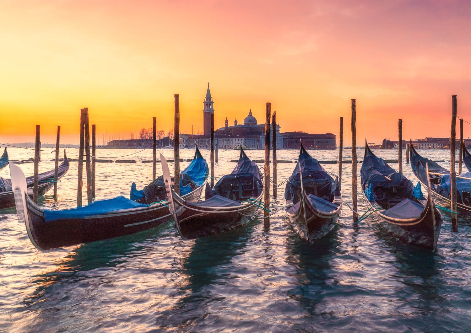 Fine art photo of gondolas floating at sunrise in Venice, with warm light on the water and San Giorgio Maggiore in the background.
