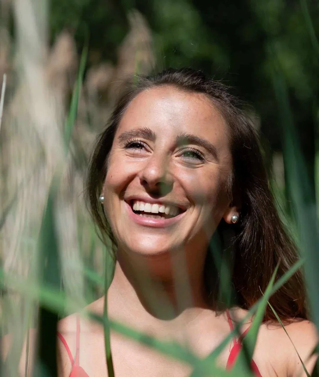 Une femme souriante avec de longs cheveux bruns, portant des boucles d'oreilles en perle, se trouve dans un champ de verdure avec des plantes hautes.