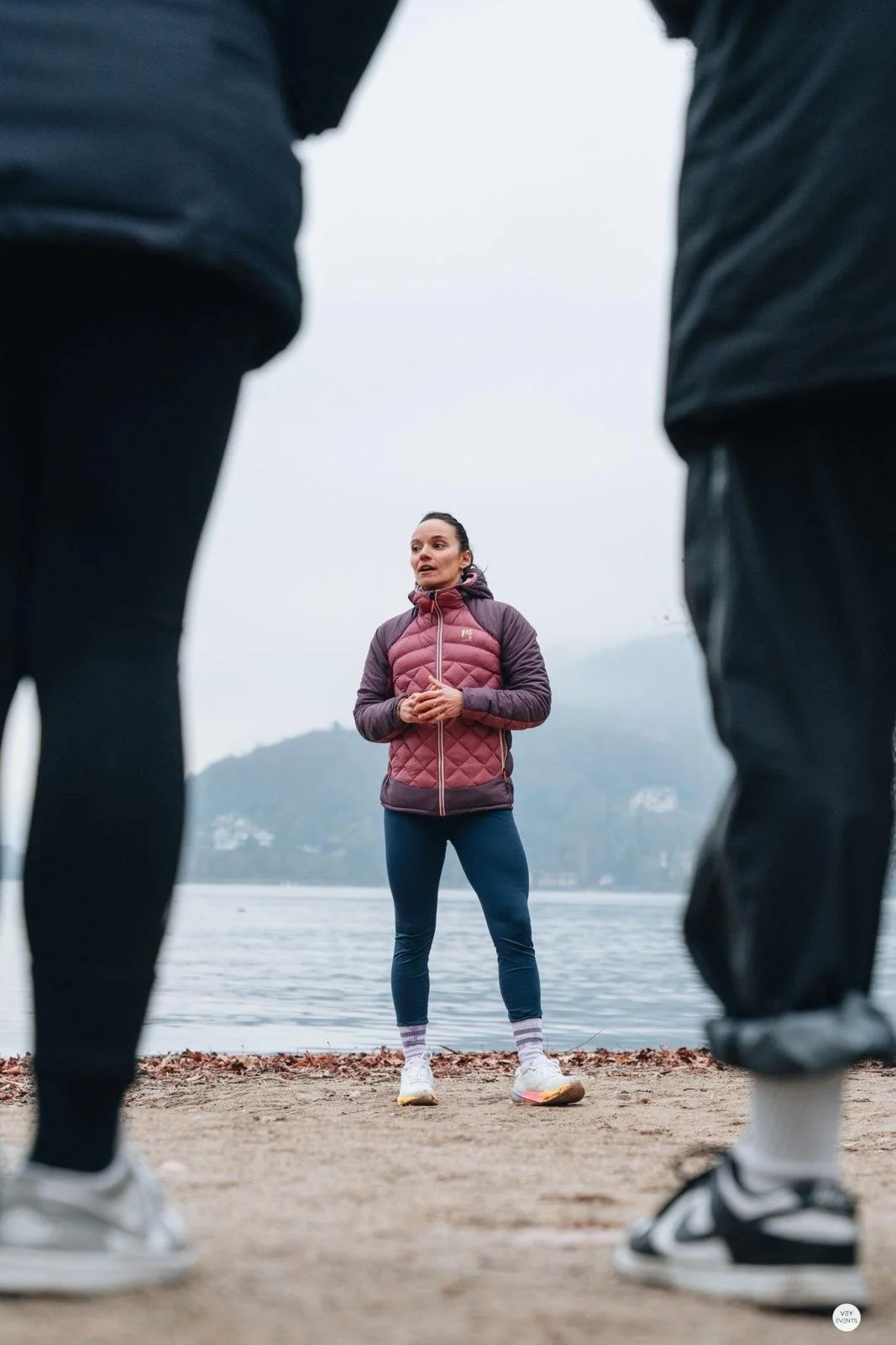 Une femme en vestiaire de sport parle à deux personnes lors d'une séance d'entraînement en extérieur au bord de l'eau dans un environnement montagneux.
