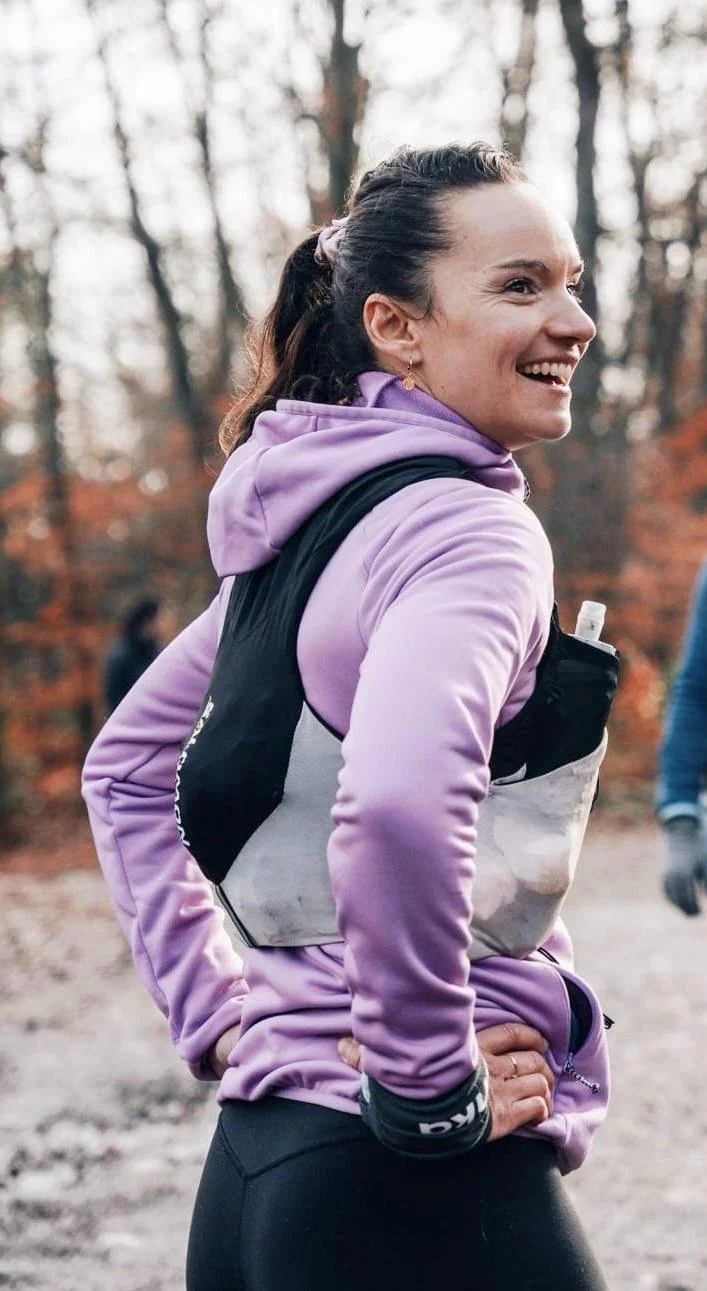 Une femme souriante portant une veste violette, un sac à dos et un gilet roulant dans une forêt en automne.
