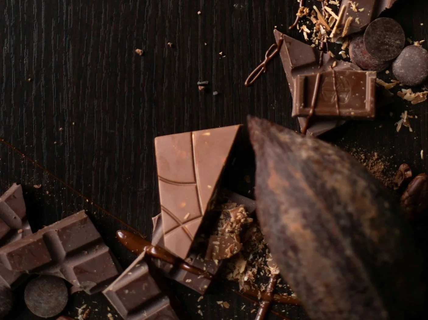 Several shattered chocolate bars and a cocoa pod rest on top of a dark wooden surface.