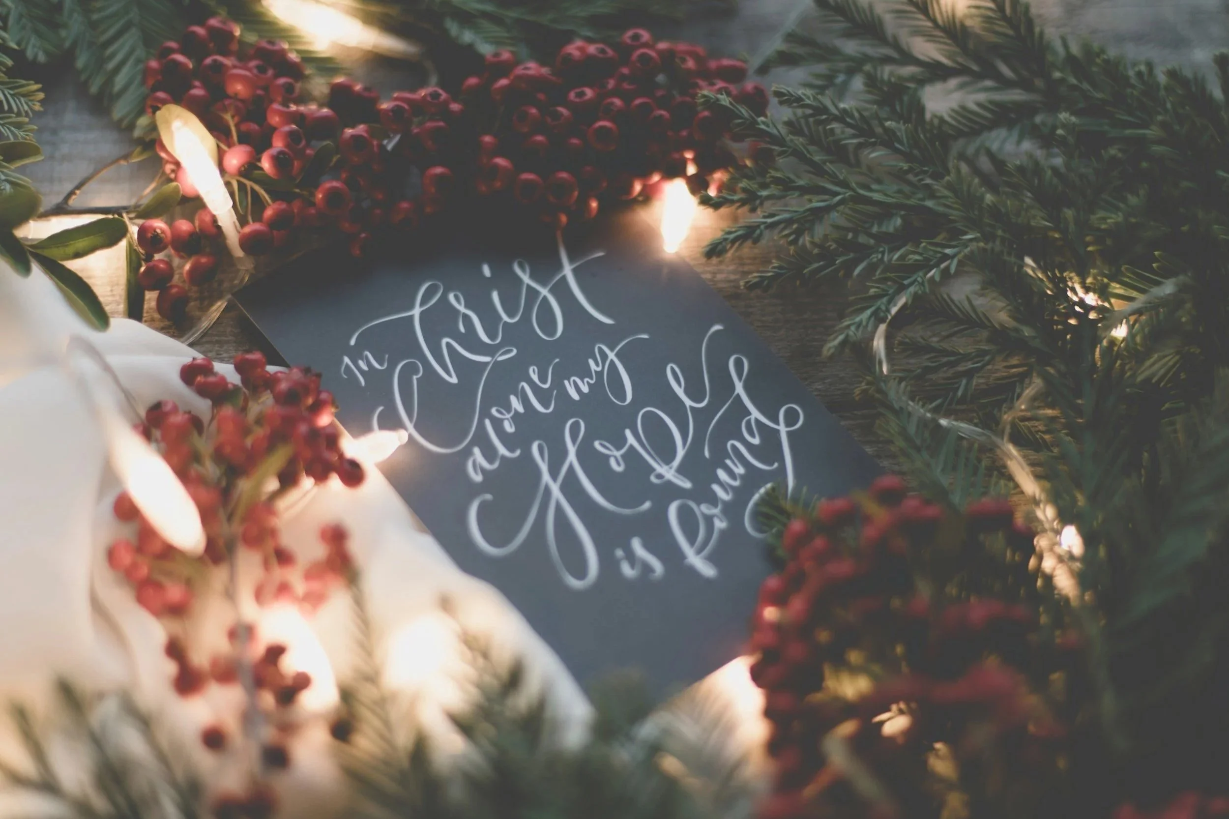 White Christmas lights and festive garlands surround a black piece of paper with white calligraphy on it. The message reads 'In Christ alone my hope is found.'
