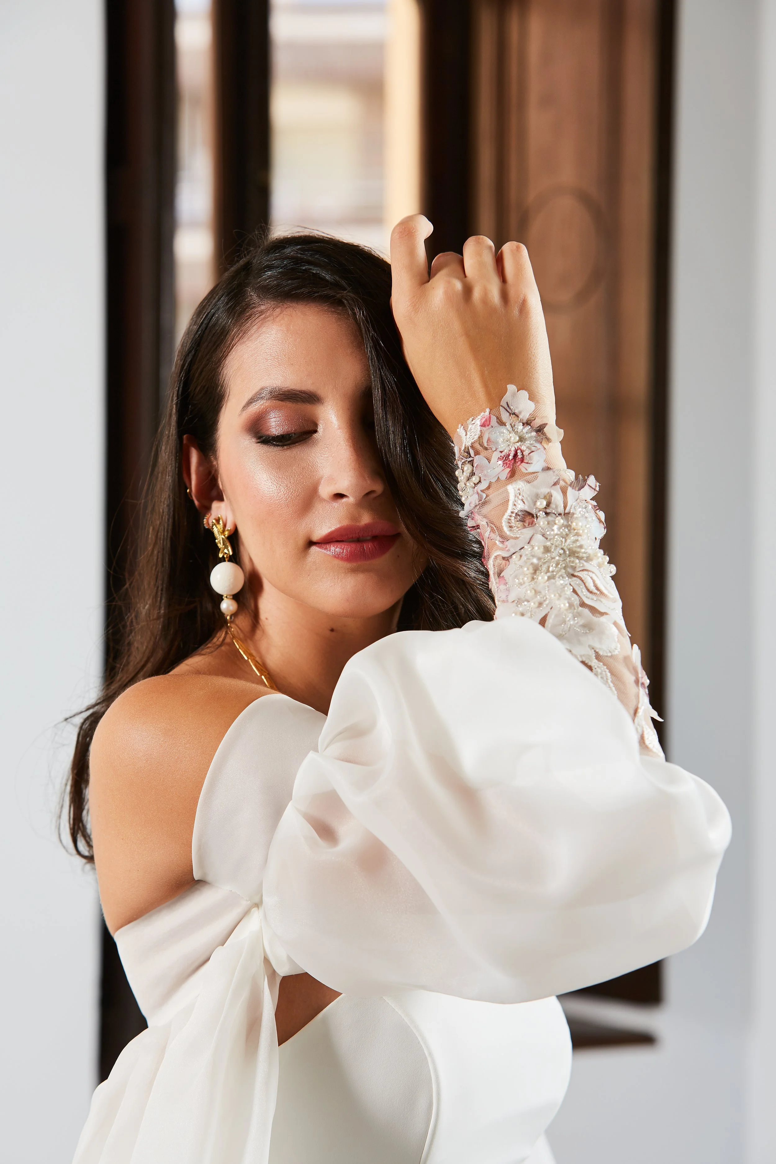 A woman with dark hair and earrings, wearing a white dress with embroidered lace sleeves, is touching her forehead with her right hand, standing near a window.