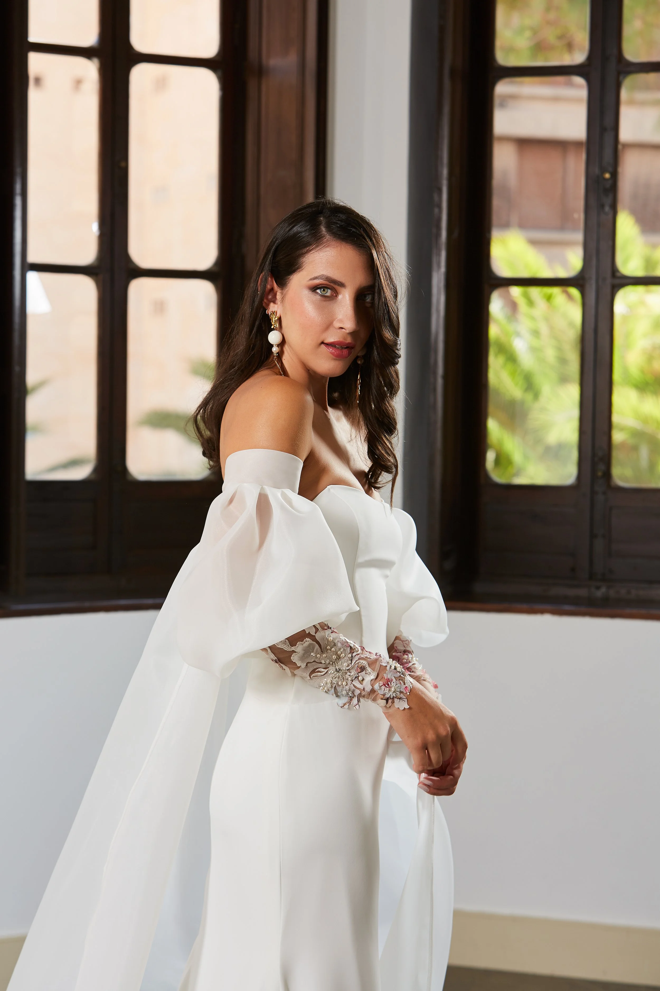 Woman in white wedding dress with off-the-shoulder puff sleeves and floral lace gloves, standing indoors near windows with greenery outside.