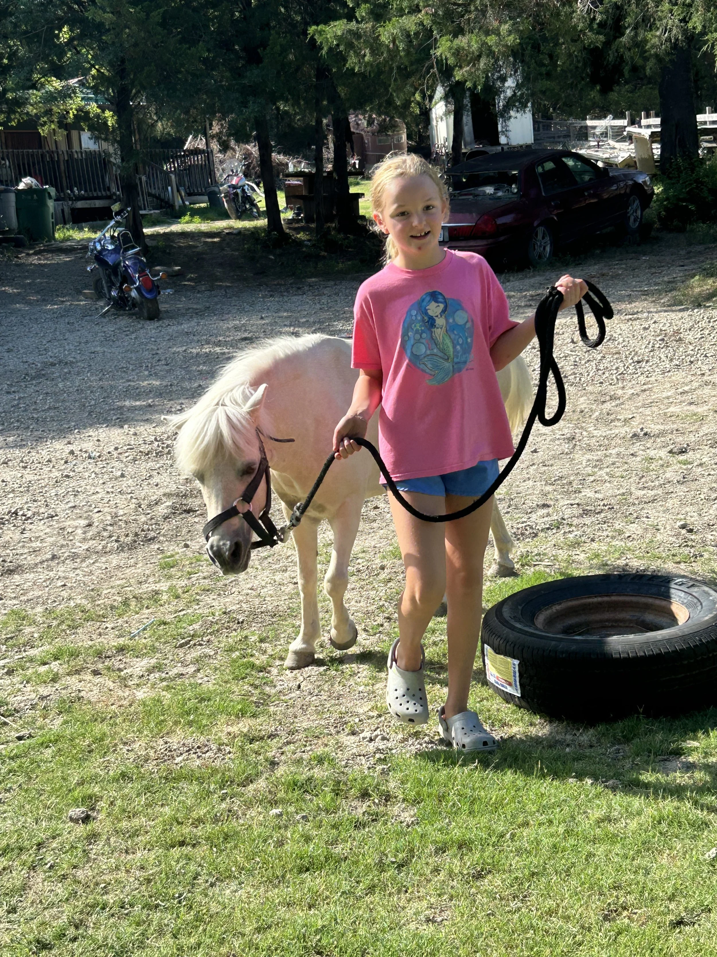 A young girl with blonde hair, wearing a pink t-shirt with a mermaid graphic, blue shorts, and white Crocs, is smiling while walking a small white pony on a black lead rope. The pony is standing near a discarded car tire on an outdoor grassy and grav