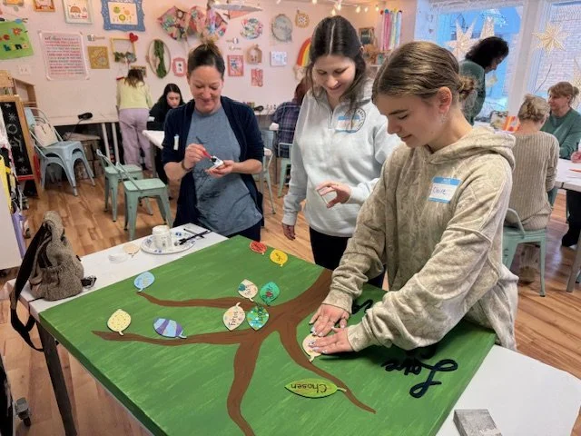 Three women working together on a large homemade tree craft project at a table in a classroom or craft room, with other people and colorful decorations in the background.